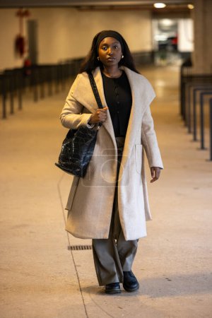 A woman walks confidently indoors in a chic beige coat, carrying a black handbag and wearing casual attire.