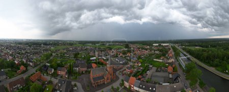 Cette photographie aérienne panoramique capture l'église Saint-Jozef à Rijkevorsel, Anvers, Belgique, sous un ciel dramatique et orageux. La vue grand-angle comprend toute la ville, mettant en valeur la