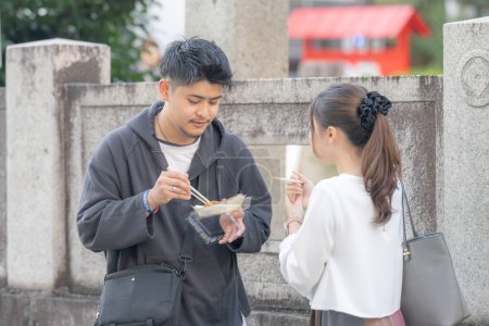  A young Japanese male and female couple in their 20s eating street food takoyaki in the back alley of Nagoya City, Aichi Prefecture