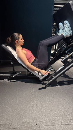 Witness the dedication of a teen woman as she focuses on training her legs in the gym. Engaged in a lower body workout session, she utilizes the press machine to strengthen and tone her leg muscles, demonstrating commitment to her fitness goals