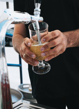 Dieses Bild zeigt einen Barkeeper, der ein erfrischendes Glas Bier aus einem Zapfhahn gießt. Die Hände der Barkeeper sind ruhig, während das goldene Bier ins Glas fließt und einen schäumenden Kopf erzeugt. Die Nahsicht