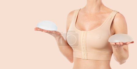Woman wearing a compressing bra after breast augmentation surgery and holding implants in hands. Copy space