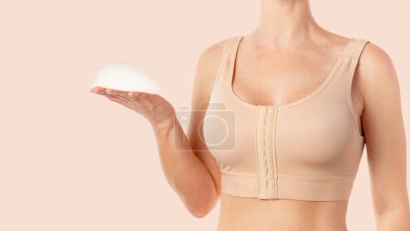 Woman wearing a compressing bra after breast augmentation surgery and holding implant in hand. Copy space