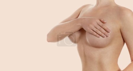 Téléchargez les photos : Dame seins nus sur fond beige couvrant la poitrine avec la main après l'augmentation mammaire. Espace de copie. - en image libre de droit
