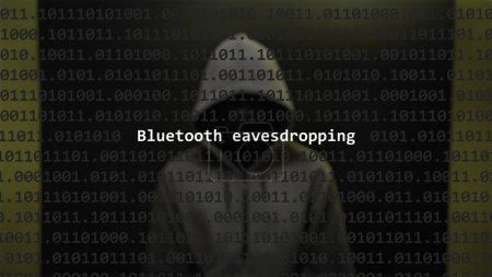 Ciberataque bluetooth escuchas texto en pantalla de primer plano, hacker anónimo oculto con capucha en el fondo borroso. Texto de vulnerabilidad en código binario del sistema en el programa editor.
