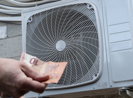 A heat pump, in foreground a hand holds out a 10 euros bill