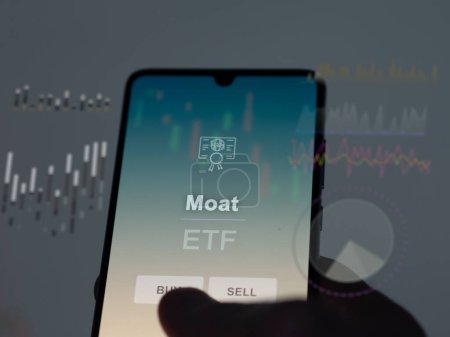 Téléchargez les photos : Un investisseur qui analyse le fonds Moat etf sur un écran. Un téléphone montre les prix des douves - en image libre de droit