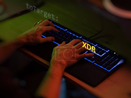 Cyber attack xdr text in foreground screen, hands of a anonymous hacker on a led keyboard. Vulnerability text in informatic system style, code on editor screen. Text in English, English text