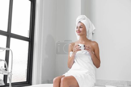Téléchargez les photos : Une femme amoureuse de beauté portant une serviette avec un corps sain et lisse, buvant du café dans une baignoire blanche dans une salle de bain avec une grande fenêtre. - en image libre de droit