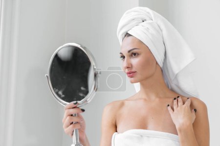 Téléchargez les photos : Une femme amoureuse de beauté portant une serviette avec un corps sain et lisse regardant un miroir rond dans une baignoire blanche dans une salle de bain. - en image libre de droit