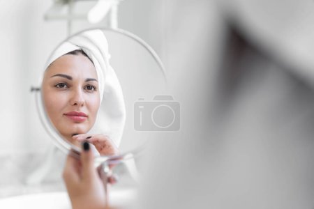 Foto de Una mujer amante de la belleza que lleva una toalla con un cuerpo sano y suave mirando un espejo redondo en una bañera blanca en un baño. - Imagen libre de derechos