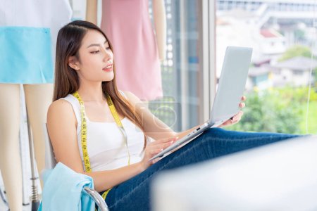 Téléchargez les photos : Les créatrices asiatiques de vêtements en Thaïlande communiquent avec les clients par le biais d'ordinateurs portables dans leur bureau à domicile. pour couper la robe selon la demande du client - en image libre de droit