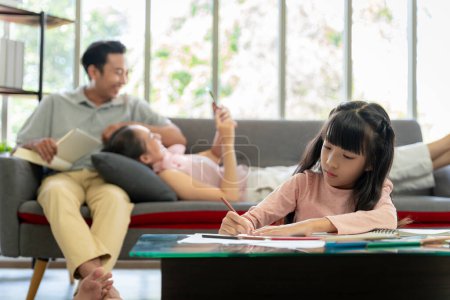Photo for An Asian girl is doing her own homework at home with her parents supervising. - Royalty Free Image
