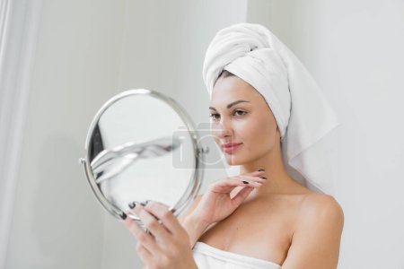 Téléchargez les photos : A beauty-loving woman wearing a towel with a healthy, smooth body looking at a round mirror in a white bathtub in a bathroom. - en image libre de droit