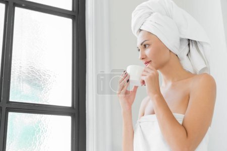 Téléchargez les photos : Une femme amoureuse de beauté portant une serviette avec un corps sain et lisse, buvant du café dans une baignoire blanche dans une salle de bain avec une grande fenêtre. - en image libre de droit
