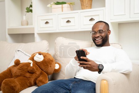 Téléchargez les photos : Africain jouer avec un téléphone intelligent Sur le canapé à la maison relaxant en vacances - en image libre de droit