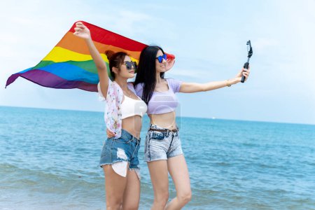 Foto de Pareja lesbiana con banderas LGBTQ en la playa, pareja feliz de vacaciones juntos en el mar - Imagen libre de derechos