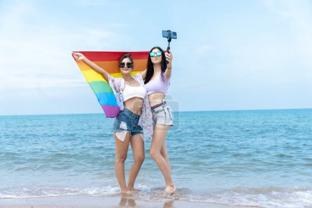 Téléchargez les photos : Couple de lesbiennes avec des drapeaux LGBTQ sur la plage, couple heureux en vacances ensemble à la mer - en image libre de droit
