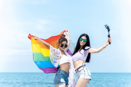 Téléchargez les photos : Couple de lesbiennes avec des drapeaux LGBTQ sur la plage, couple heureux en vacances ensemble à la mer - en image libre de droit