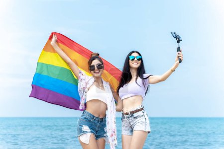 Foto de Pareja lesbiana con banderas LGBTQ en la playa, pareja feliz de vacaciones juntos en el mar - Imagen libre de derechos