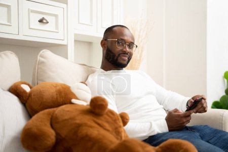 Foto de Africano jugando con el teléfono inteligente En el sofá en casa relajándose de vacaciones - Imagen libre de derechos