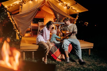 Glückliche Familie entspannen und verbringen Zeit zusammen in Glamping an Sommerabenden und Gitarre spielen am gemütlichen Lagerfeuer. Luxus-Zeltlager für Erholung und Erholung im Freien. Lifestyle-Konzept