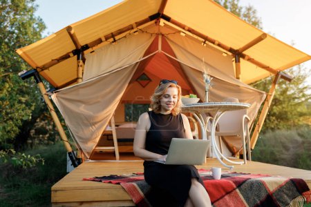 Foto de Happy Woman freelancer using a laptop on a cozy glamping tent in a sunny day. Luxury camping tent for outdoor summer holiday and vacation. Lifestyle concept - Imagen libre de derechos