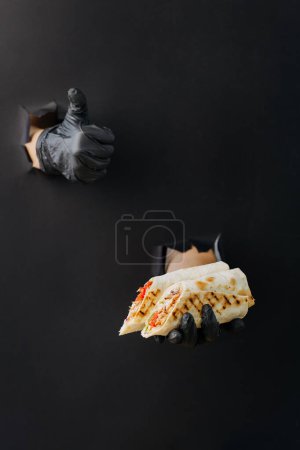 Téléchargez les photos : Main tenant donateur ou shawarma et montrant un pouce à travers des trous déchirés dans fond de papier noir - en image libre de droit