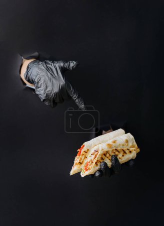 Téléchargez les photos : Main tenant donateur ou shawarma à travers des trous déchirés dans fond de papier noir. Concept de nourriture de rue - en image libre de droit