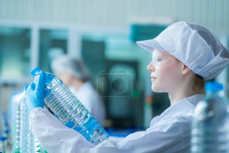 Téléchargez les photos : Travailleur scientifique de vérifier la qualité des bouteilles d'eau sur la ligne de convoyeur de la machine à l'usine industrielle. Travailleuses enregistrant les données de la chaîne de production de boissons. - en image libre de droit