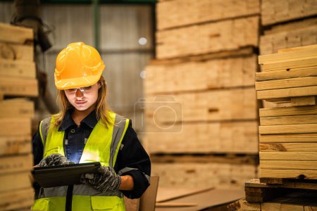 Téléchargez les photos : Ouvrière charpentier portant un uniforme de sécurité et un casque dur travaillant et vérifiant la qualité des produits en bois lors de la fabrication en atelier. hommes et femmes travailleurs bois dans l'industrie des entrepôts sombres. - en image libre de droit