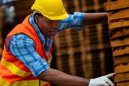 Téléchargez les photos : Travailleur charpentier africain portant un uniforme de sécurité et un casque dur travaillant et vérifiant la qualité des produits en bois lors de la fabrication en atelier. hommes et femmes travailleurs bois dans l'industrie des entrepôts sombres. - en image libre de droit