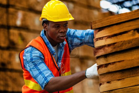 Foto de Carpintero trabajador africano con uniforme de seguridad y sombrero duro trabajando y comprobando la calidad de los productos de madera en la fabricación del taller. hombres y mujeres trabajadores madera en la industria de almacenes oscuros. - Imagen libre de derechos