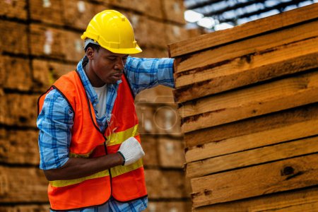 Foto de Carpintero trabajador africano con uniforme de seguridad y sombrero duro trabajando y comprobando la calidad de los productos de madera en la fabricación del taller. hombres y mujeres trabajadores madera en la industria de almacenes oscuros. - Imagen libre de derechos