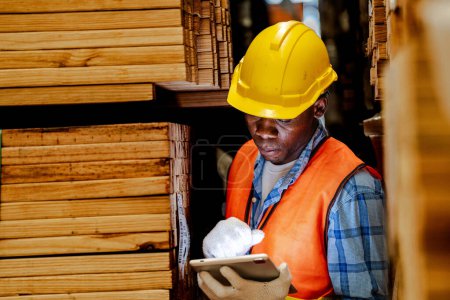Téléchargez les photos : Travailleur charpentier africain portant un uniforme de sécurité et un casque dur travaillant et vérifiant la qualité des produits en bois lors de la fabrication en atelier. hommes et femmes travailleurs bois dans l'industrie des entrepôts sombres. - en image libre de droit