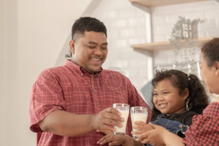 Photo for An asian family are drinking milk. They were happily inviting their girl to drink morning milk together in the kitchen of their home. Breakfast time of Asian dad mom and kid. - Royalty Free Image