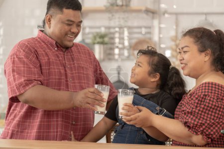 Photo for An asian family are drinking milk. They were happily inviting their girl to drink morning milk together in the kitchen of their home. Breakfast time of Asian dad mom and kid. - Royalty Free Image