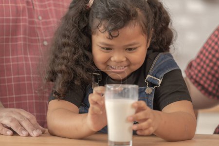 Téléchargez les photos : Fille tient un verre de lait. Ils étaient heureux d'inviter leur fille à boire du lait le matin ensemble dans la cuisine de leur maison. Petit déjeuner de papa asiatique maman et les enfants. - en image libre de droit
