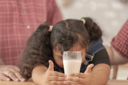 Photo for Girl is holding a glass of milk. They were happily inviting their girl to drink morning milk together in the kitchen of their home. Breakfast time of Asian dad mom and kid people. - Royalty Free Image