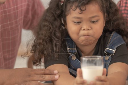 Photo for Girl is holding a glass of milk. They were happily inviting their girl to drink morning milk together in the kitchen of their home. Breakfast time of Asian dad mom and kid people. - Royalty Free Image