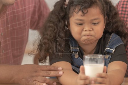 Téléchargez les photos : Fille tient un verre de lait. Ils étaient heureux d'inviter leur fille à boire du lait le matin ensemble dans la cuisine de leur maison. Petit déjeuner de papa asiatique maman et les enfants. - en image libre de droit