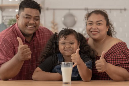 Photo for Girl is smiling with glass of milk. They were happily inviting their girl to drink morning milk together in the kitchen of their home. Breakfast time of Asian dad mom and kid people. - Royalty Free Image