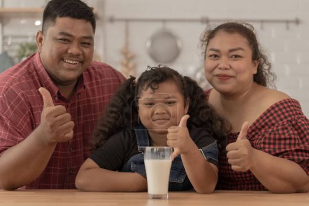 Photo for Girl is smiling with glass of milk. They were happily inviting their girl to drink morning milk together in the kitchen of their home. Breakfast time of Asian dad mom and kid people. - Royalty Free Image