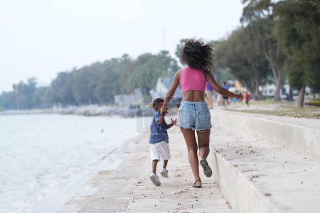 Téléchargez les photos : Mixte race africaine et asiatique mère et garçon joue à l'espace extérieur. souriant famille heureuse avoir du plaisir à courir sur la plage. portrait de maman et d'enfant style de vie avec une coiffure unique. - en image libre de droit