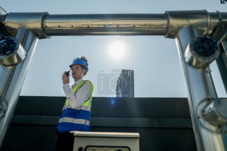 Foto de Ingeniero en equipo de seguridad lleva a cabo la inspección de grandes tanques de agua industrial en una azotea del edificio. Ingeniero profesional verifica meticulosamente la conectividad y el estado de las tuberías en una azotea urbana. - Imagen libre de derechos