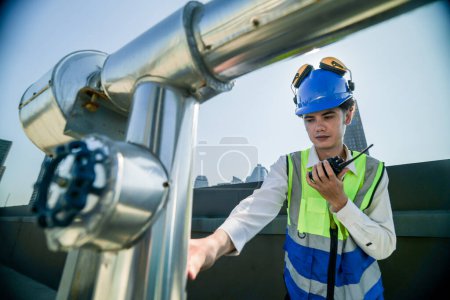 Engineer in safety gear conducts inspection on large industrial water tanks on a building rooftop. Professional engineer meticulously checks connectivity and status of piping on an urban rooftop.