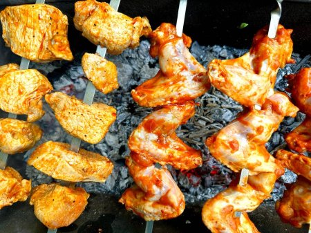 Foto de Las alitas de pollo marinadas y los kebabs en las brochetas se asan a la parrilla, dando vuelta lentamente para freír. Carne kebab en llamas. Asar carne. Cocinar carne en un picnic - Imagen libre de derechos