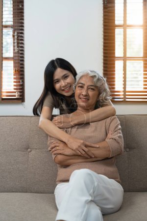 Liebevolle erwachsene Tochter umarmt ältere Mutter auf Couch zu Hause, Familie genießt zärtlichen Moment zusammen, junge Frau und reife Mutter oder Großmutter schauen einander an, zwei Generationen.