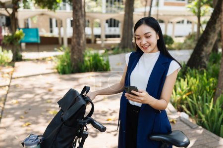 Asiatische junge Geschäftsfrau radelt mit umweltfreundlicher grüner Energie zur Arbeit und nutzt das Smartphone, während sie in der Stadt pendelt. Umweltfreundlich.