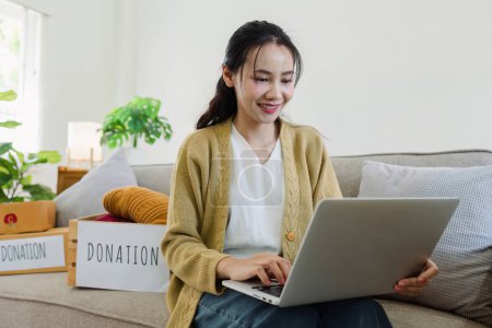 Asiatische junge Frau nutzt Laptop für Spendenbox mit Altkleidern.